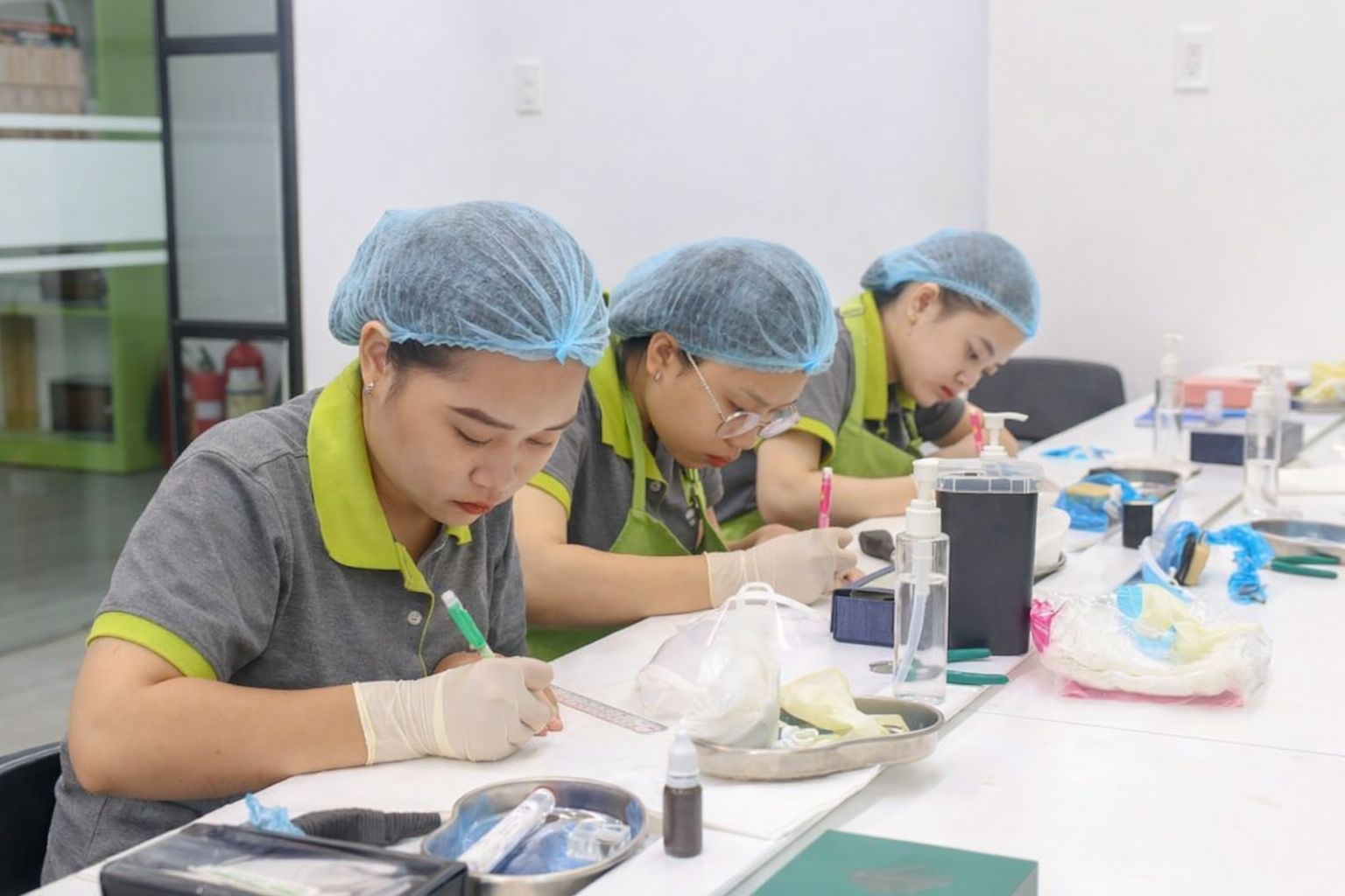 Tre kvinnor i hygienmössor och handskar övar nagelterapi vid ett vitt bord.